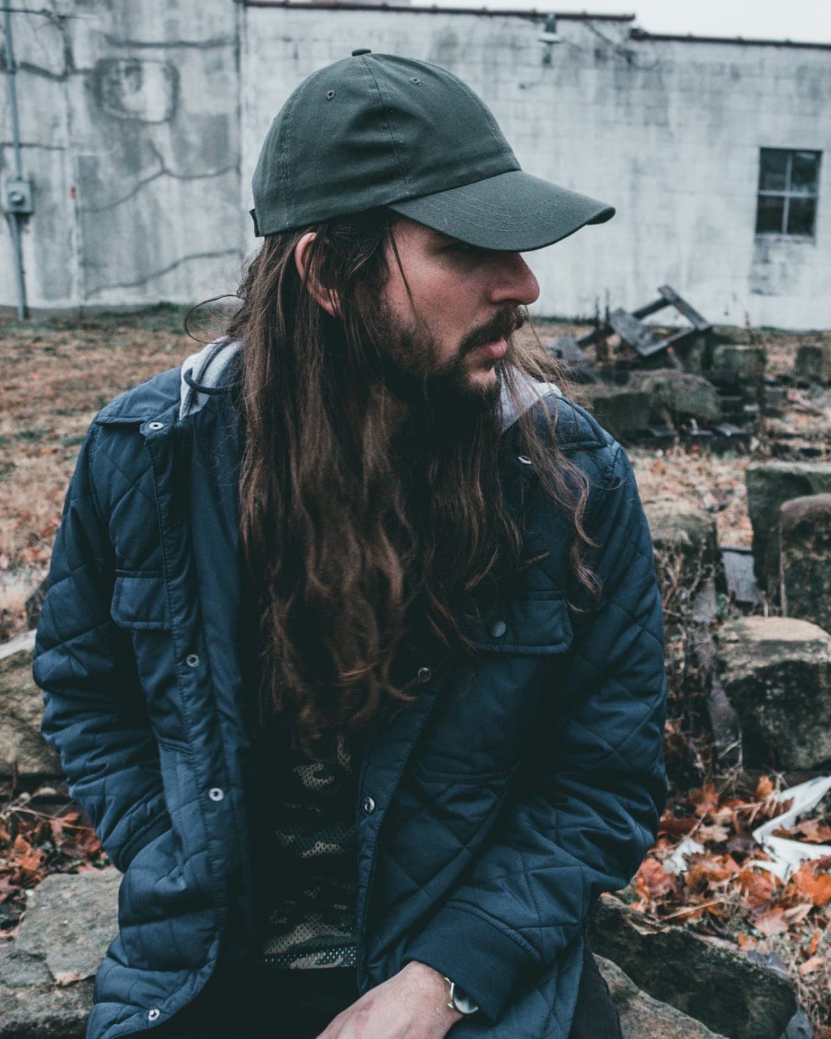 A long-haired man in a cap and a padded jacket sitting among autumn leaves