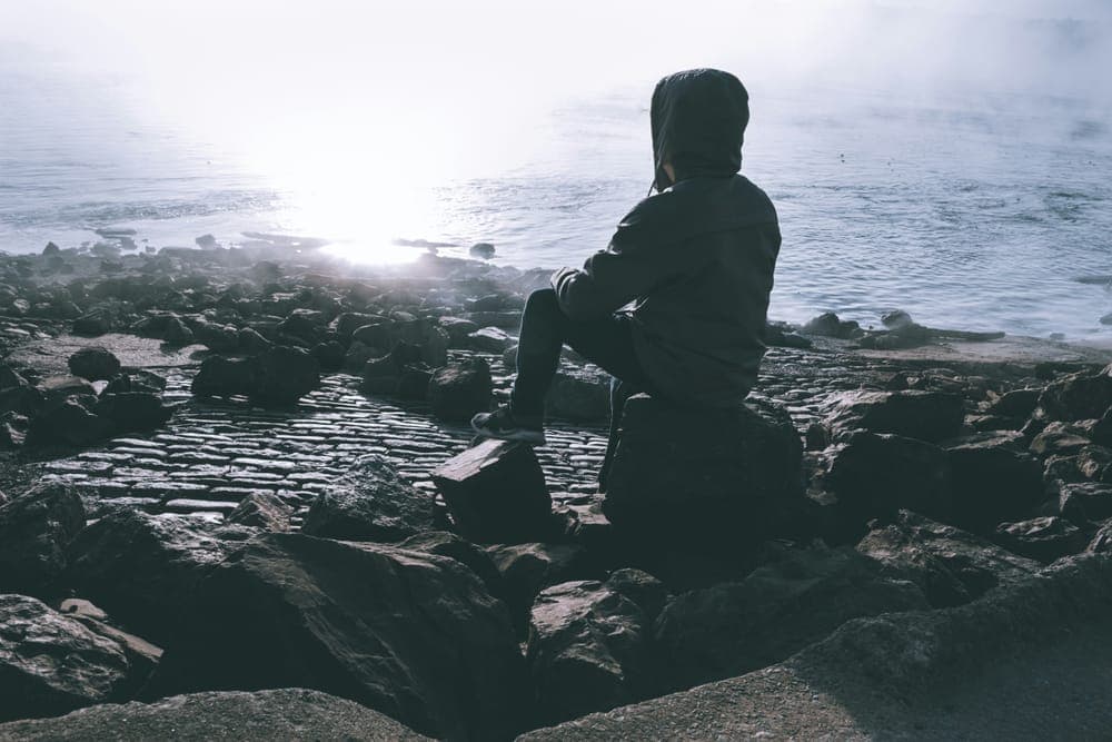 A figure in a hood sitting on dark rocks by the sea in misty light