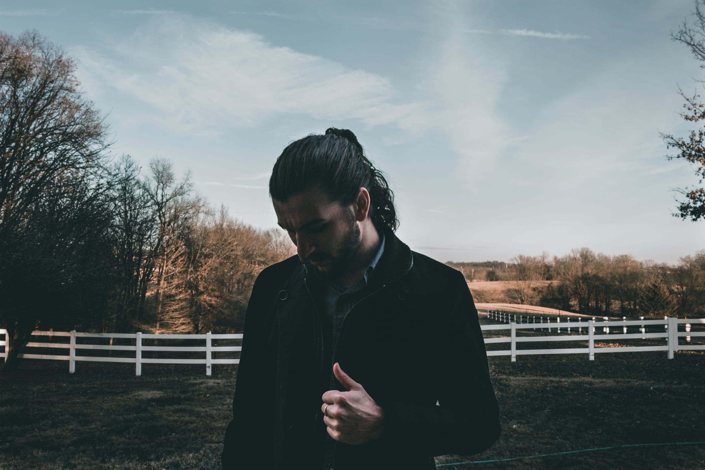 A man with a long ponytail in a dark coat, looking down, in front of a white fence and bare trees