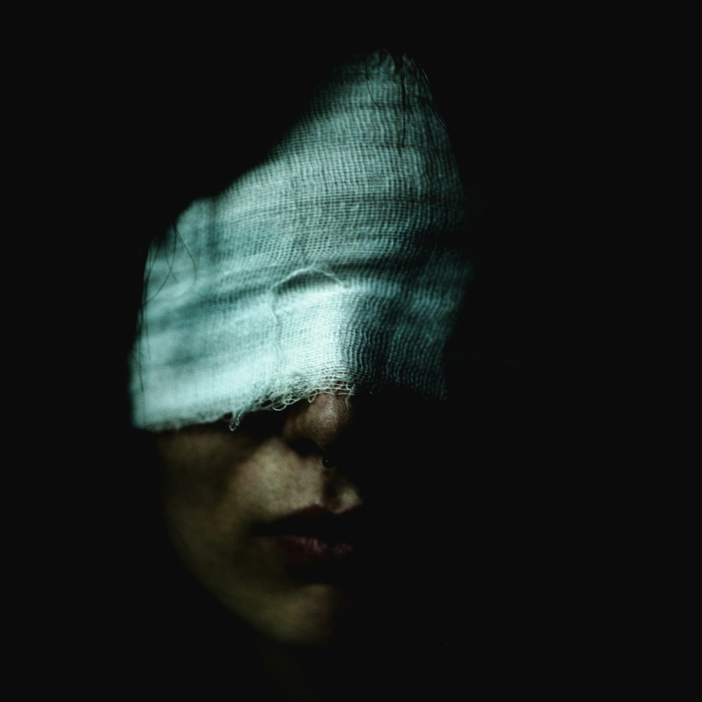 A face with a pale cloth over the eyes against a black background
