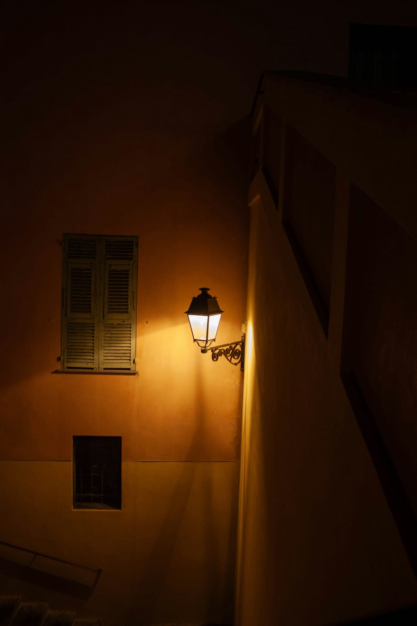 A street lamp and shuttered window on a warm-lit wall at night
