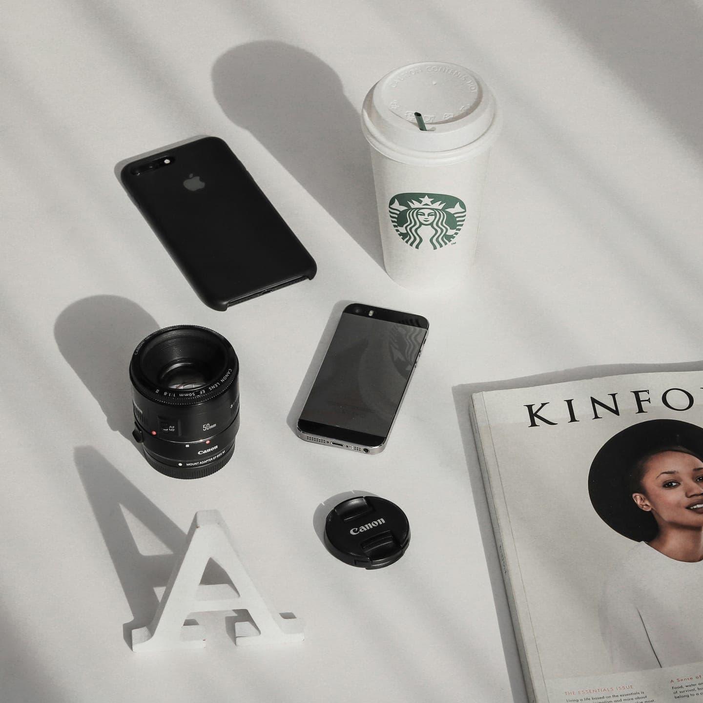 A camera lens, a phone, a coffee cup and a Kinfolk magazine on a white table, seen from above