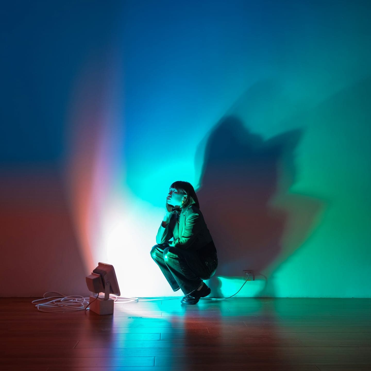 A woman crouching in a studio lit in teal and red, her shadow on the wall