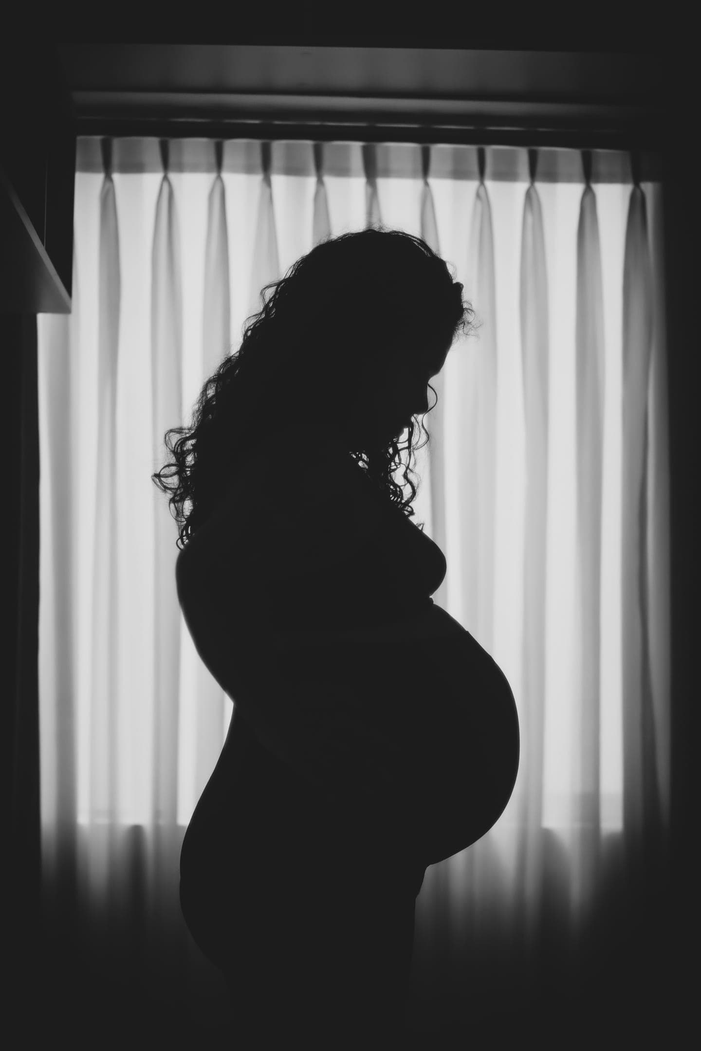 A pregnant woman in silhouette against a curtained window