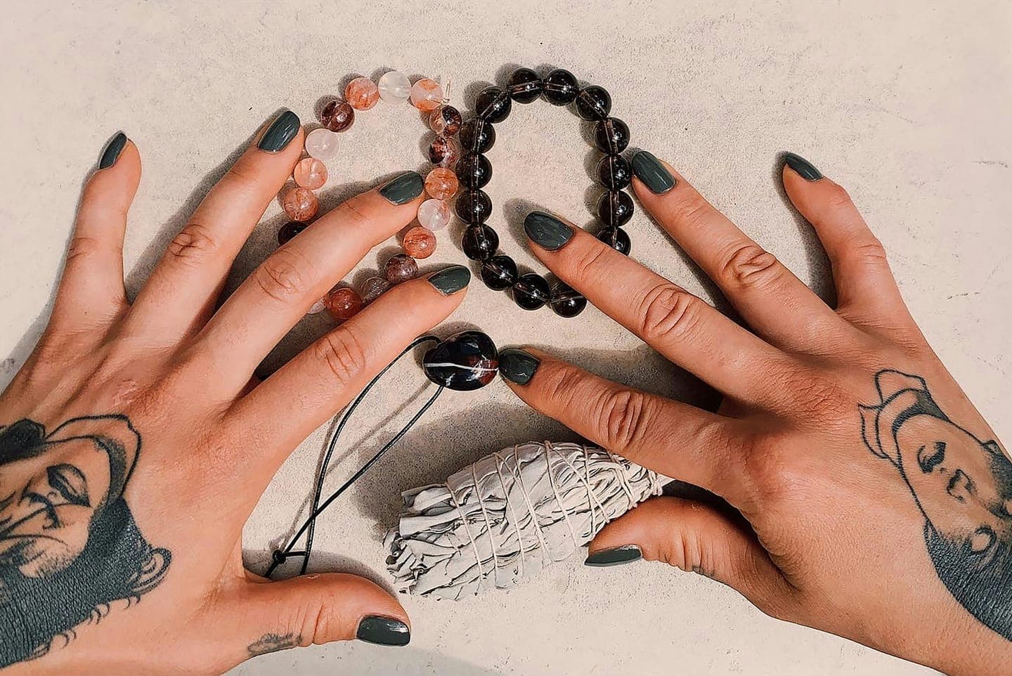Two tattooed hands with bead bracelets on a pale surface