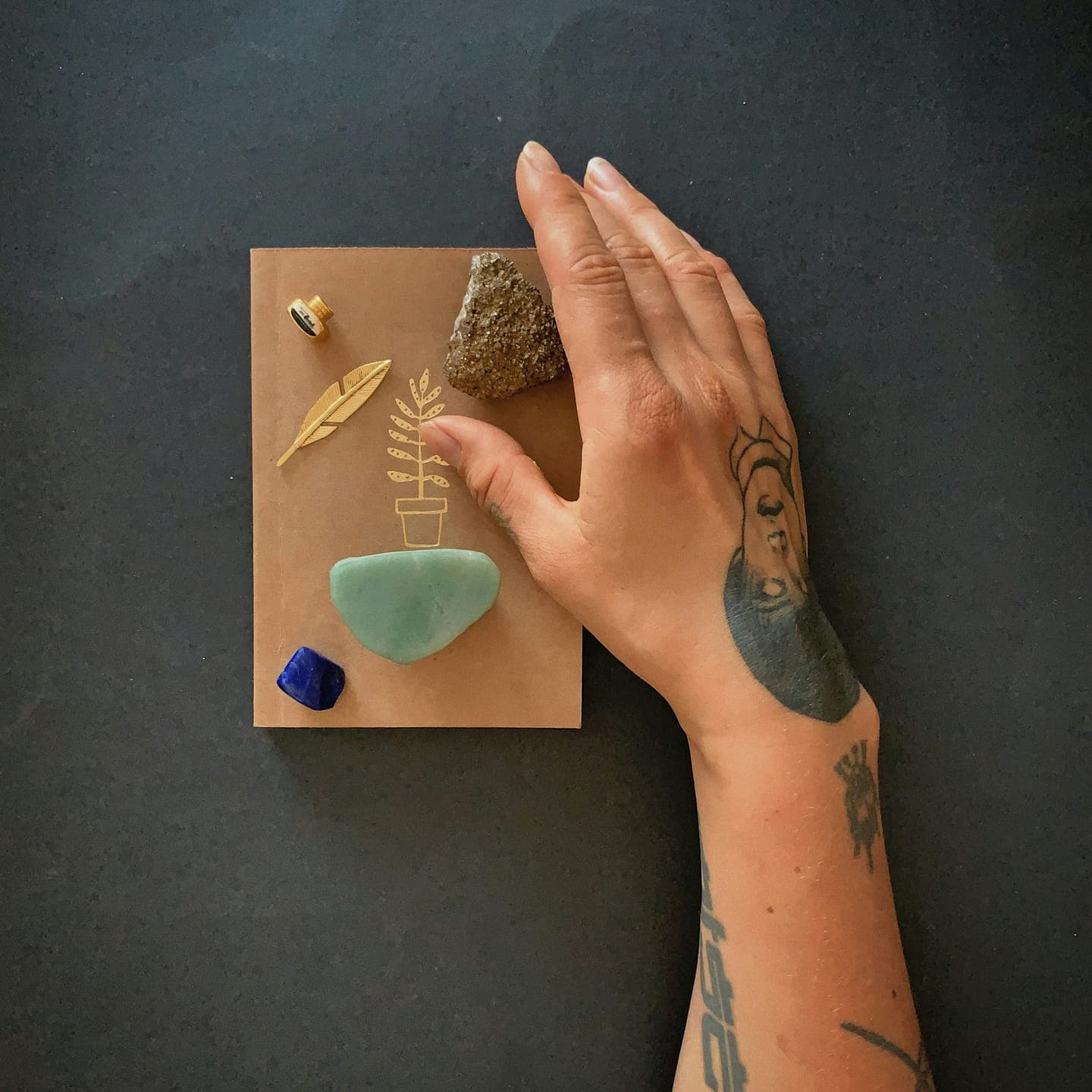 A tattooed hand arranging small stones and a leaf on a card